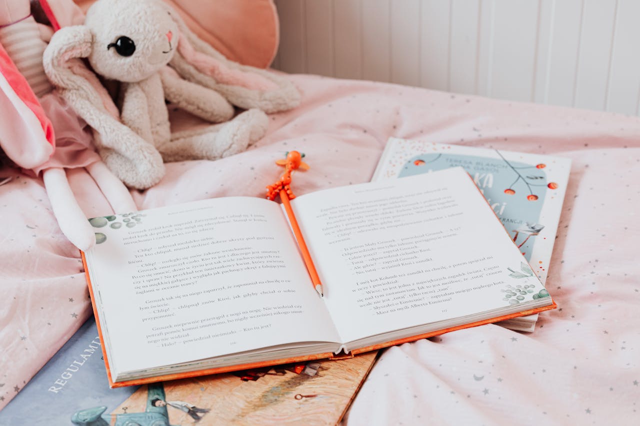 services-03 An open book with pencil on a pink bed, accompanied by a stuffed bunny toy, creating a cozy reading vibe.